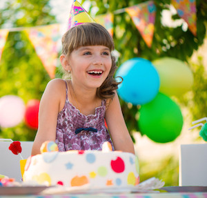 Little girl at a birthday party, laughing and sitting in front of a birthday cake with a birthday hat on and balloons in background